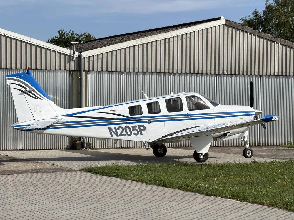 A white and blue Beechcraft Bonanza N205P parked on a paved surface with a hangar and trees in the background.