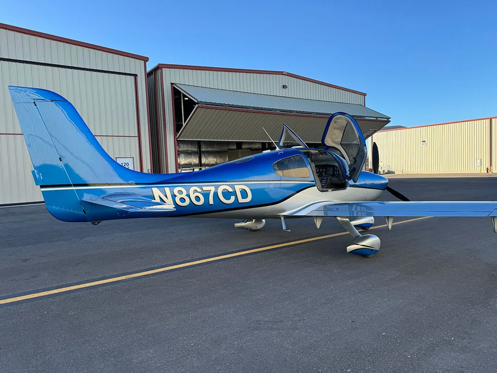 A side view of a blue and silver Cirrus SR22 aircraft with registration N867CD, parked with its doors open.