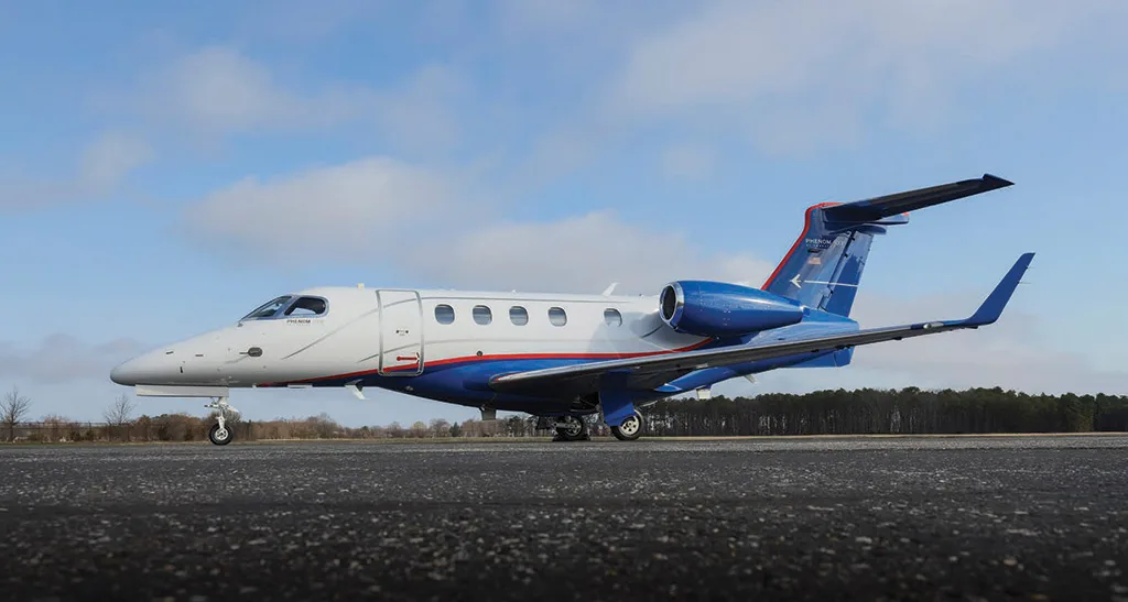 A white and blue Embraer Phenom 300 business jet is parked on an asphalt runway under a partly cloudy sky.