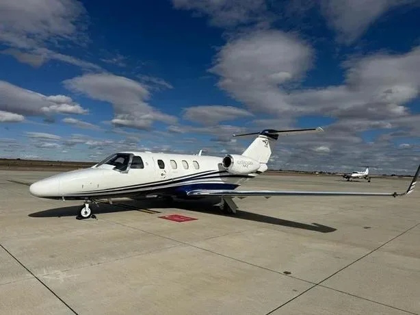 A white Cessna Citation M2 business jet with blue and black stripes parked on an airfield under a partly cloudy sky.