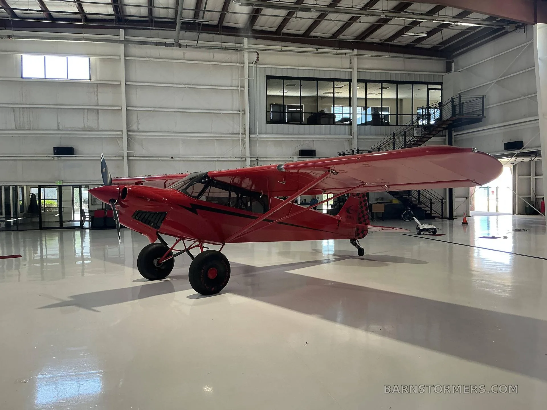A red Bushmaster aircraft with large tires is parked in a hangar, viewed from the front-left, showing the hangar interior.