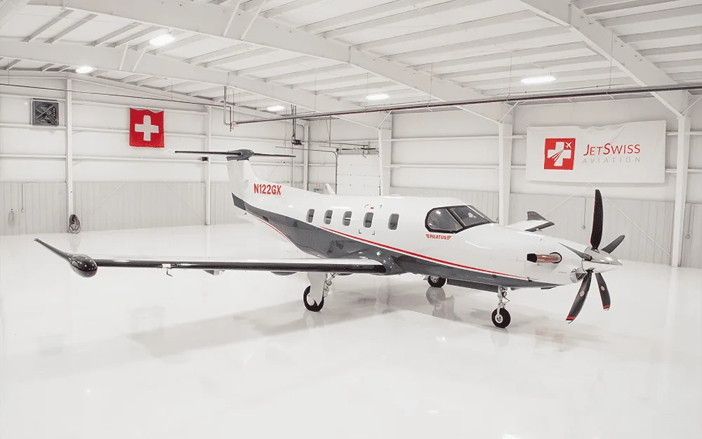 A white and grey Pilatus PC-12 turboprop aircraft with tail number N122GX parked inside a bright white hangar.