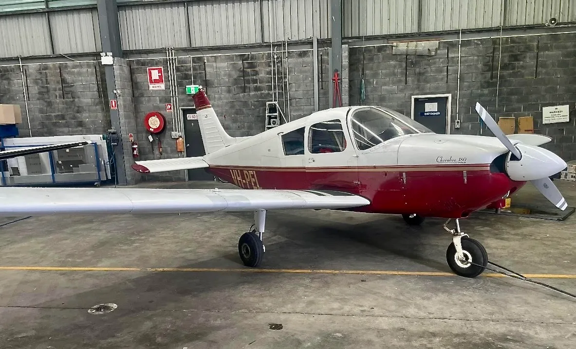 A red and white Piper Cherokee 180 aircraft, registration VH-PEL, is parked inside a hangar with a concrete block wall.