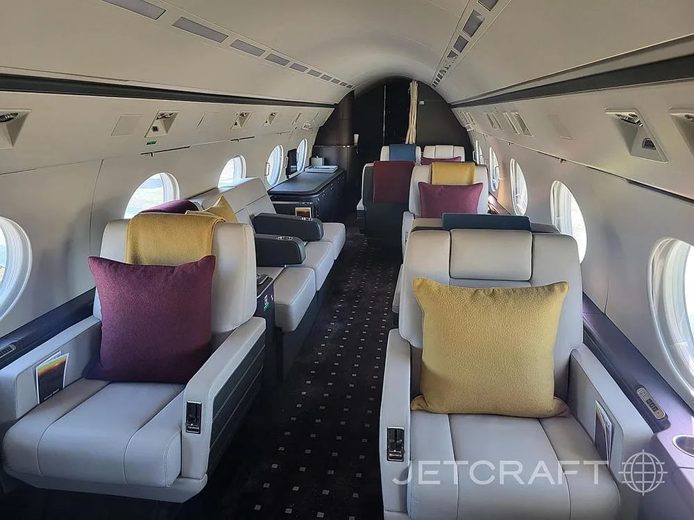 The luxurious interior cabin of a private jet, featuring light-colored seats with colorful pillows and dark patterned carpet.