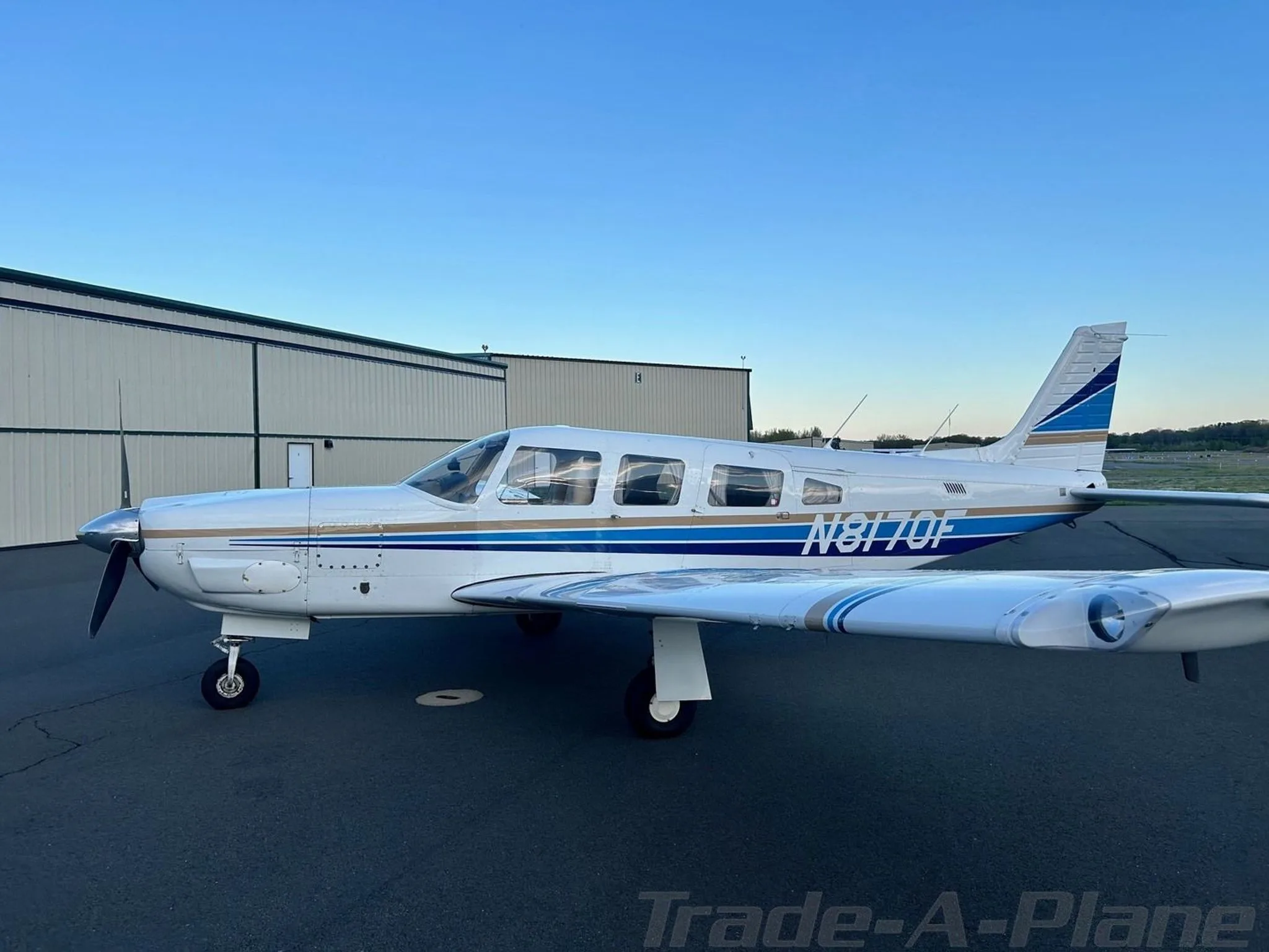 A white Piper Cherokee aircraft with blue and gold stripes, tail number N8170F, parked on an airfield next to a hangar.