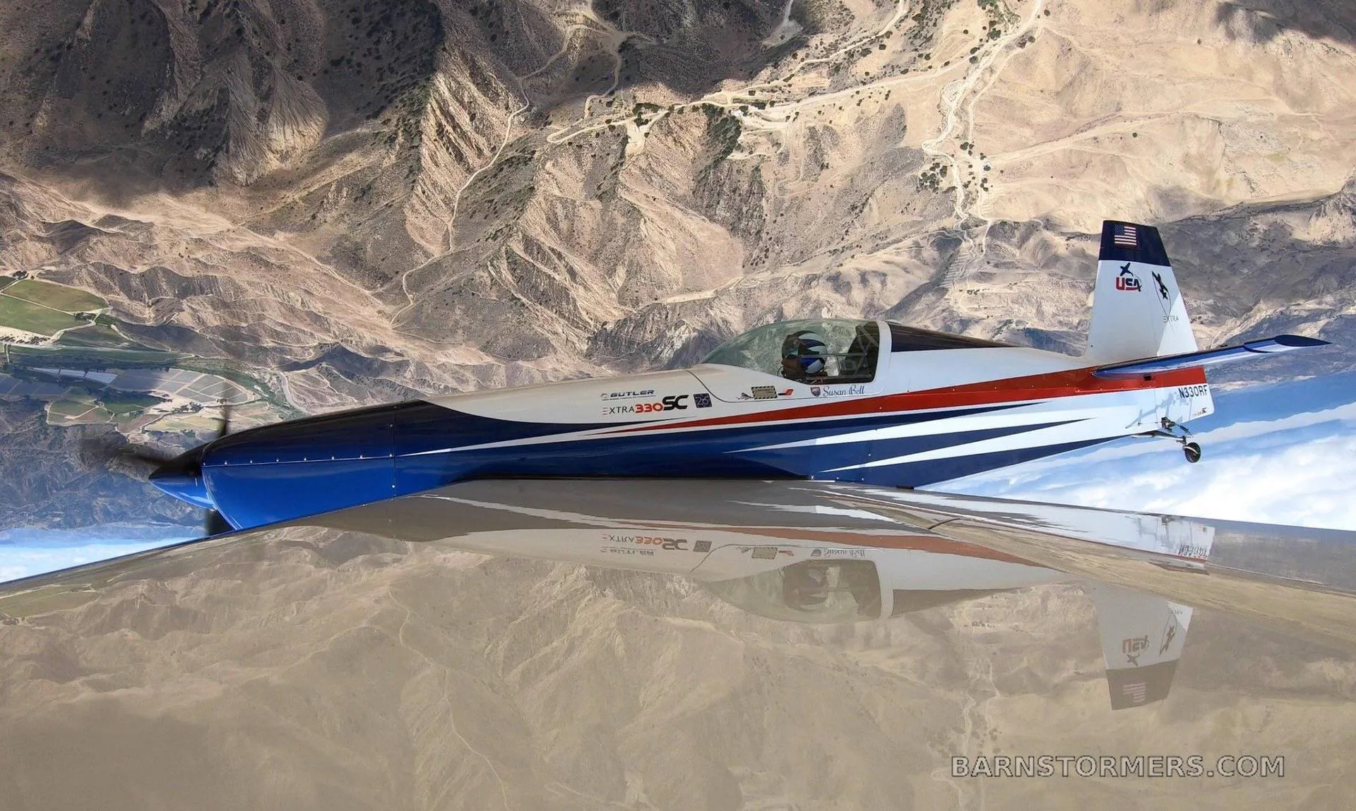 An Extra 330SC aircraft with blue, white, and red stripes flies inverted over rugged desert mountains.