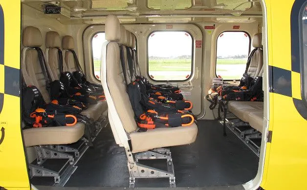 The interior cabin of a yellow and black helicopter shows rows of beige seats with orange safety harnesses.