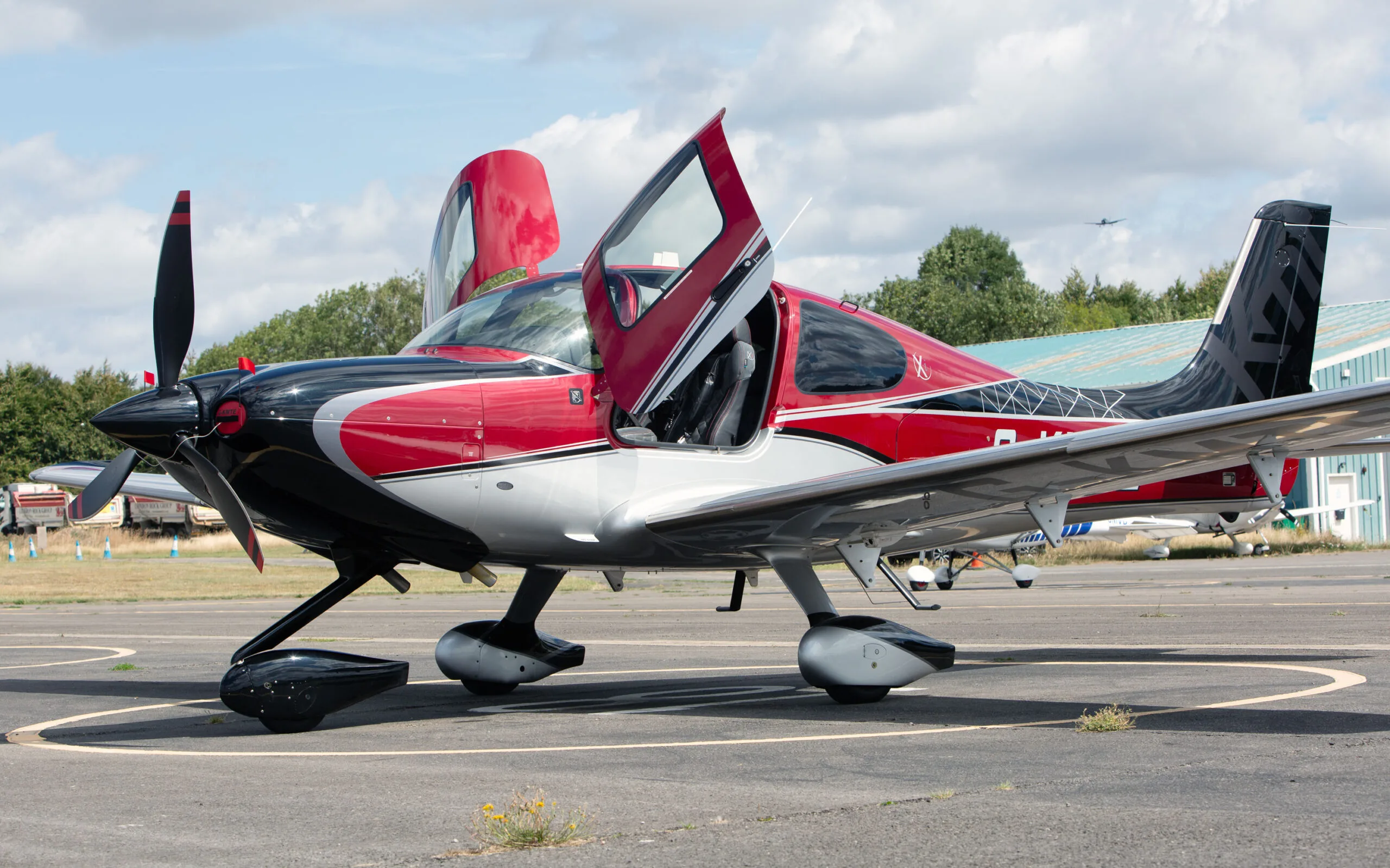 A red, black, and silver Cirrus SR22 aircraft with its gull-wing doors open, parked on an airfield.