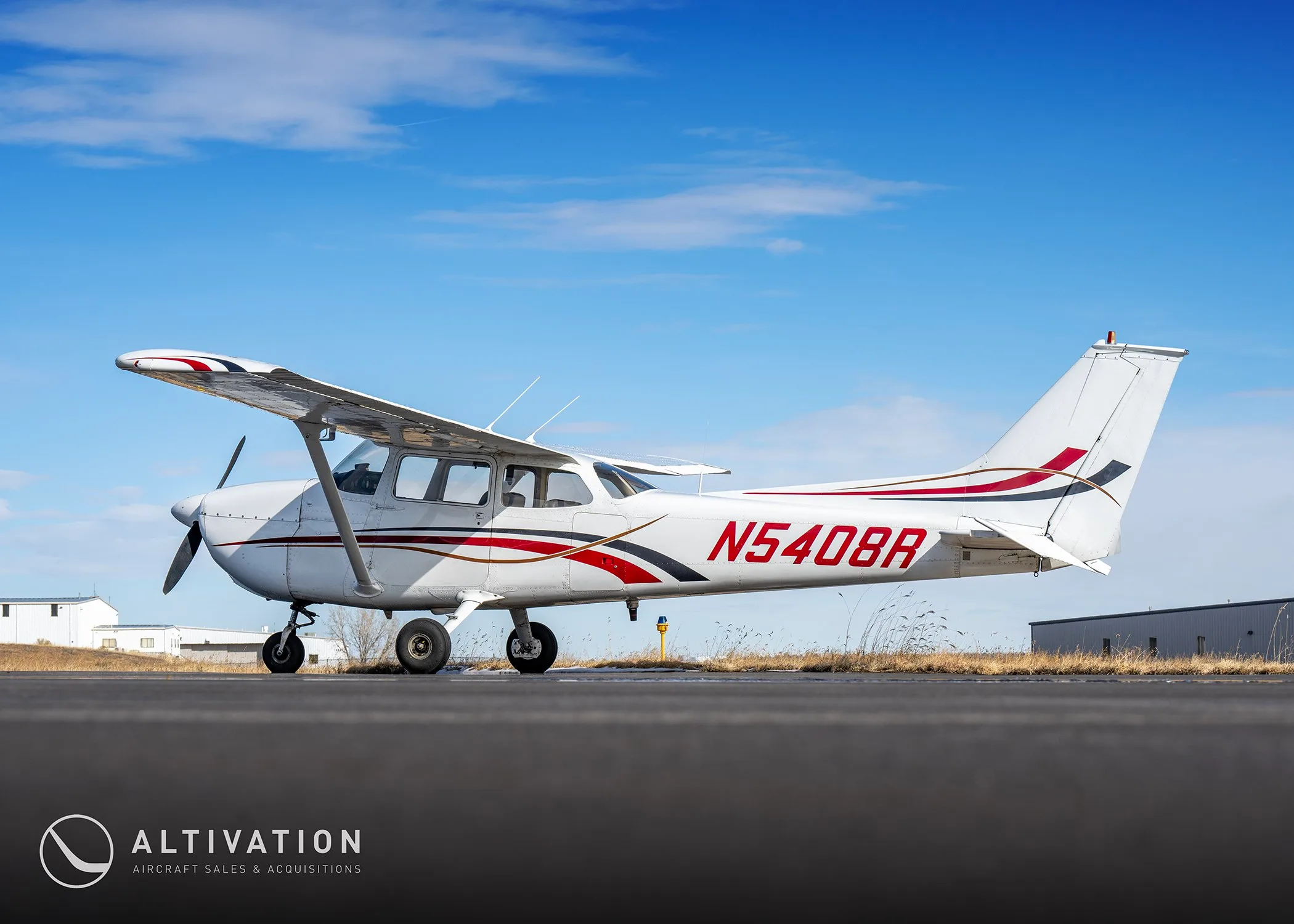A side view of a white Cessna 172 with red and black stripes, registration N5408R, parked on an airfield under a blue sky.