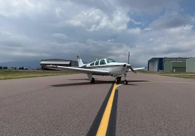 A white and gold Beechcraft Bonanza aircraft is parked on an airfield, centered on a yellow taxiway line under a cloudy sky.