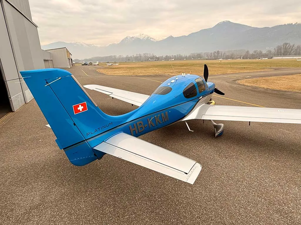 A rear-quarter view of a blue and silver Cirrus SR22T aircraft with registration HB-KKM on an airfield.