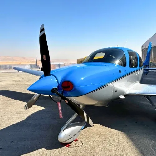 A blue and silver single-engine propeller aircraft, possibly a Cirrus, parked on a tarmac with a 'REMOVE BEFORE FLIGHT' tag.