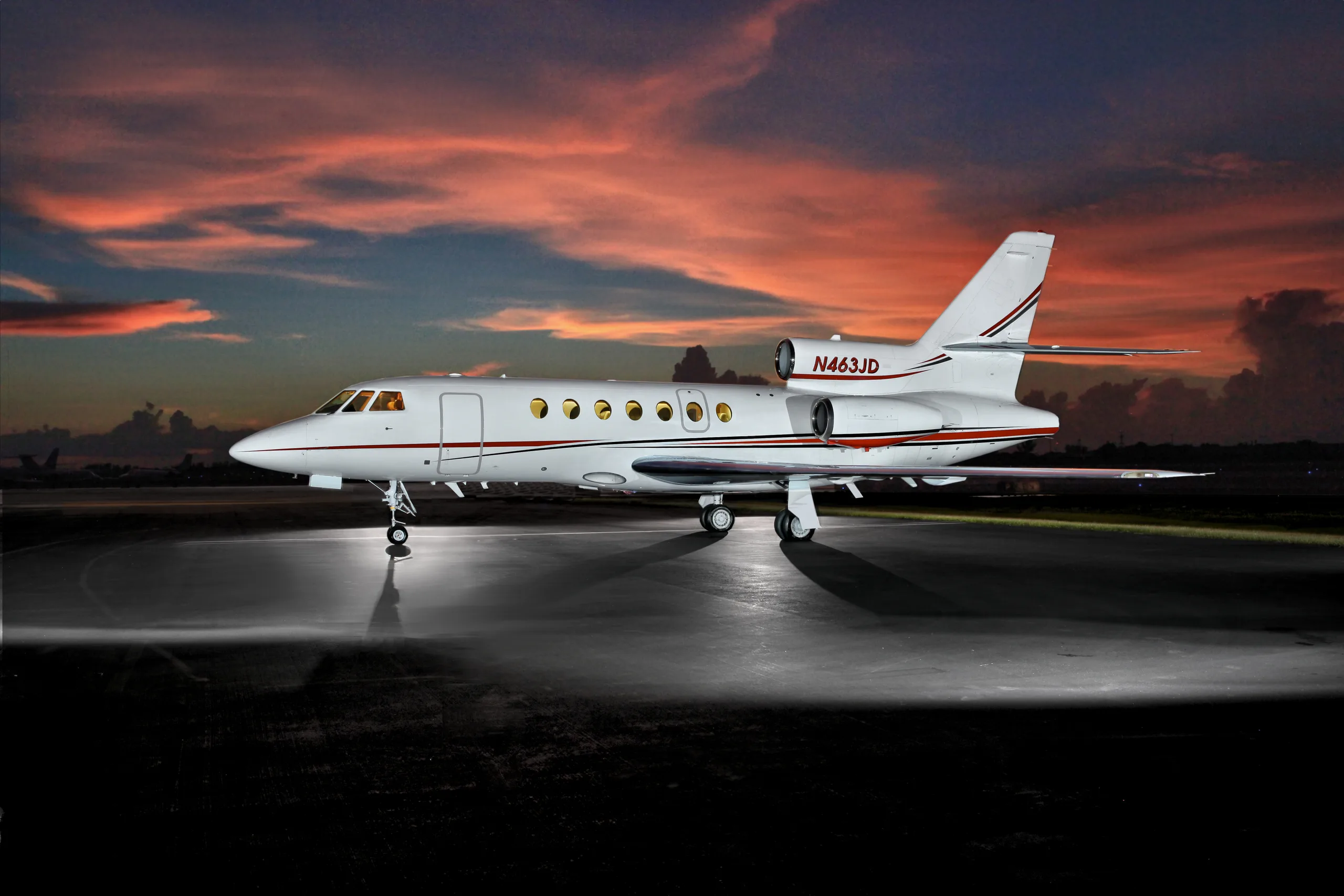 A white business jet with red and black stripes on the tail and engines is parked on an tarmac at sunset.