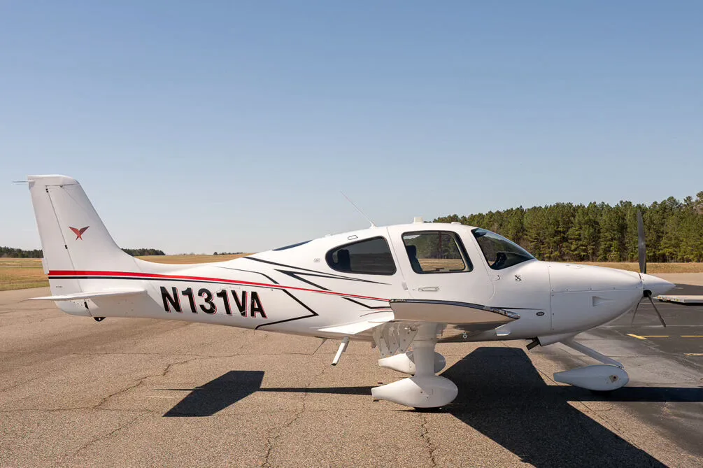 A white Cirrus SR22 aircraft with red and black accent stripes, tail number N131VA, parked on an airfield with trees in the background.