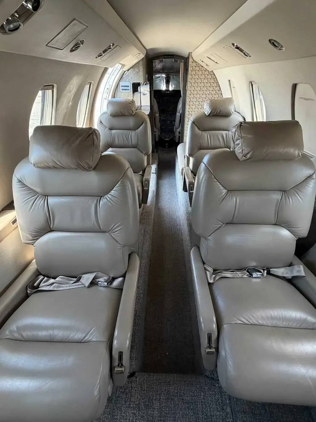 A view down the aisle of a private jet cabin, showing four beige leather seats in a club configuration.