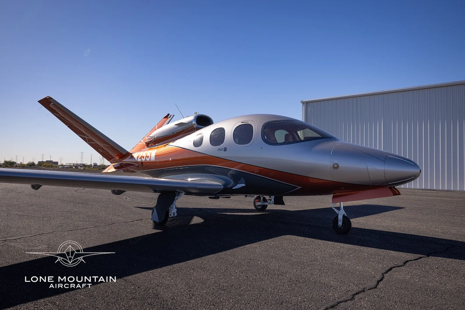A Cirrus Vision Jet (SF50) in silver and orange paint scheme, parked on an asphalt tarmac with a hangar in the background.