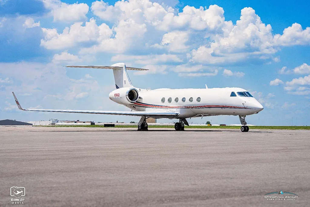 A white Gulfstream G550 aircraft with red and blue stripes on the fuselage, parked on an airfield under a partly cloudy sky.