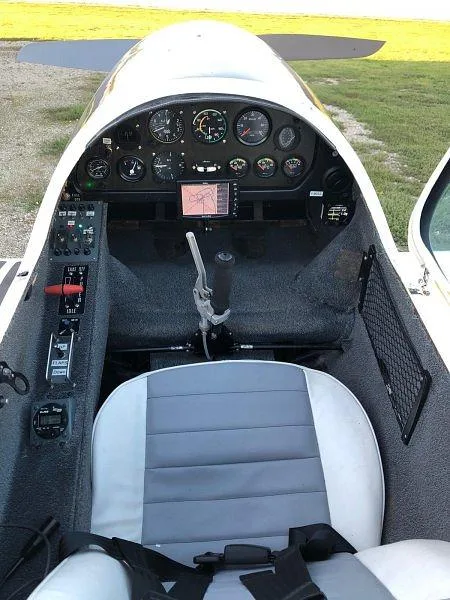 The cockpit of a small aircraft showing the instrument panel with analog gauges and a central screen, and a grey and white seat.