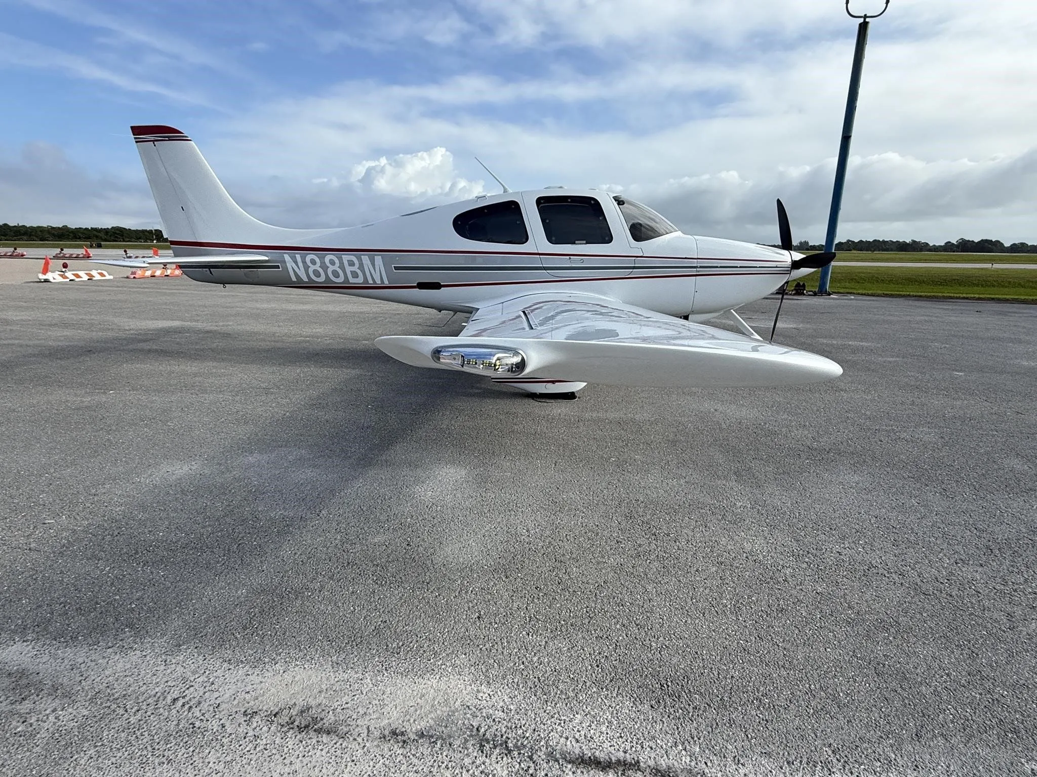 A white Cirrus SR22 aircraft with red and gray stripes, tail number N88BM, parked on an airport tarmac, showing the right wing and landing gear.
