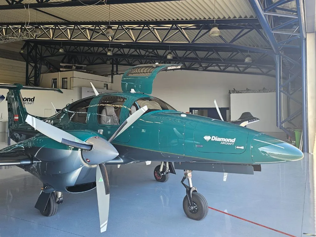 A dark green Diamond DA62 aircraft with its doors open is parked inside a hangar.