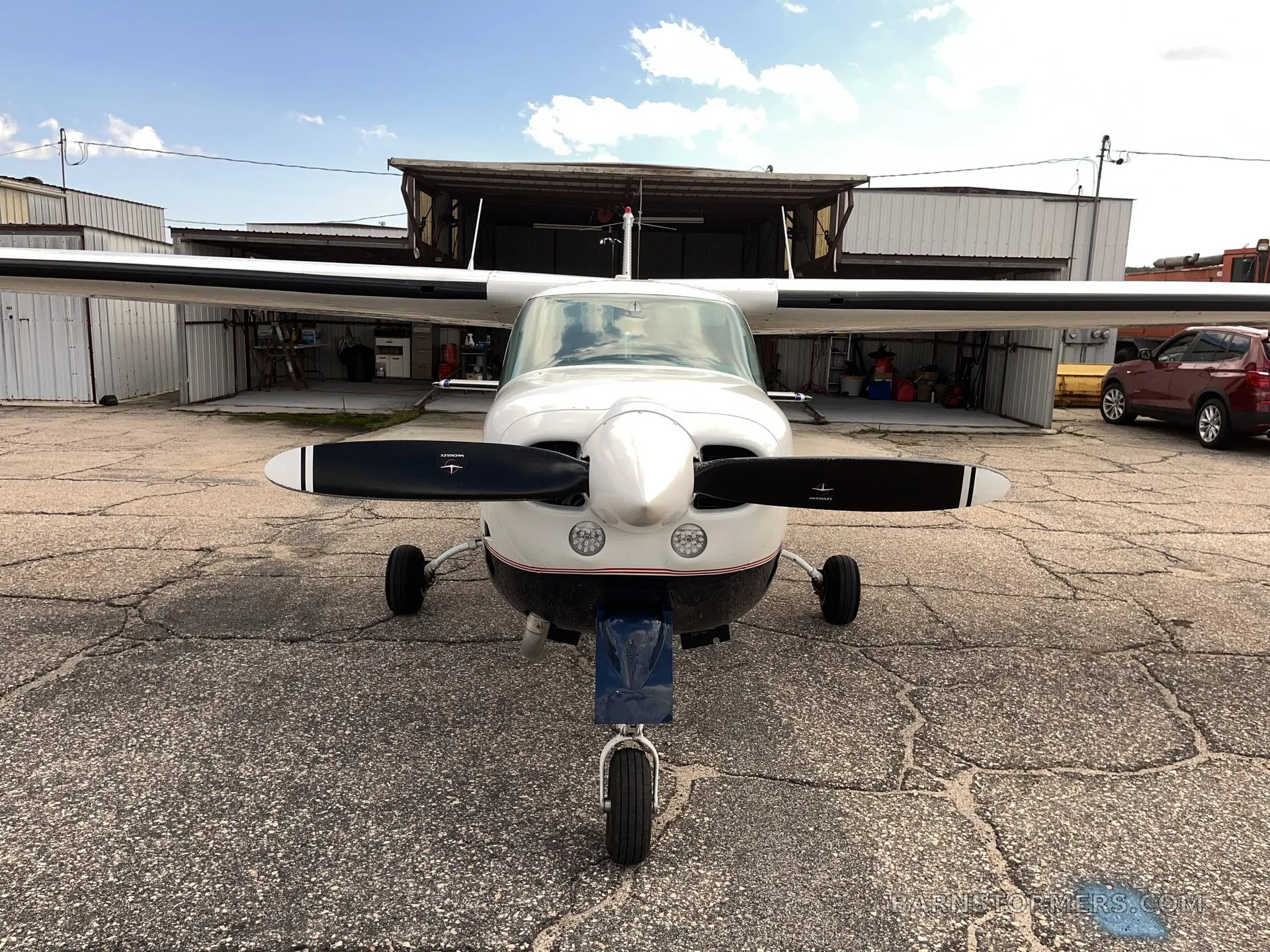 Front view of a white and dark blue single-engine propeller aircraft with a two-blade propeller, parked on a concrete apron in front of a hangar.