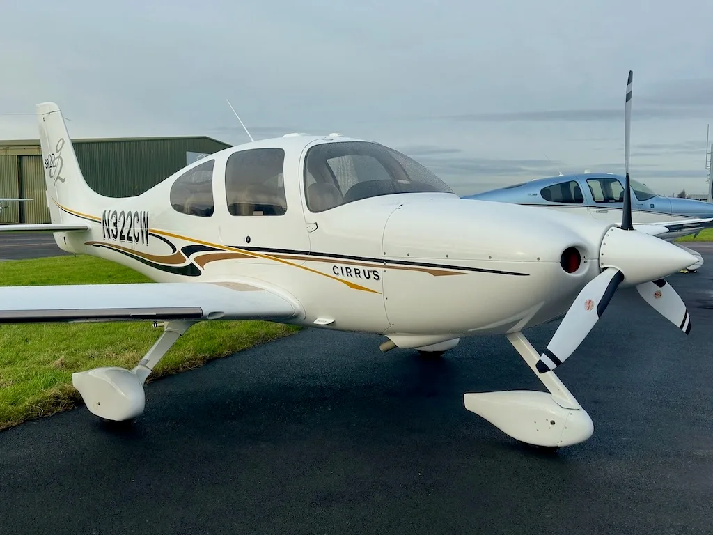A white Cirrus SR22 aircraft with N322CW registration and gold and black stripes, parked on a tarmac with a hangar in the background.