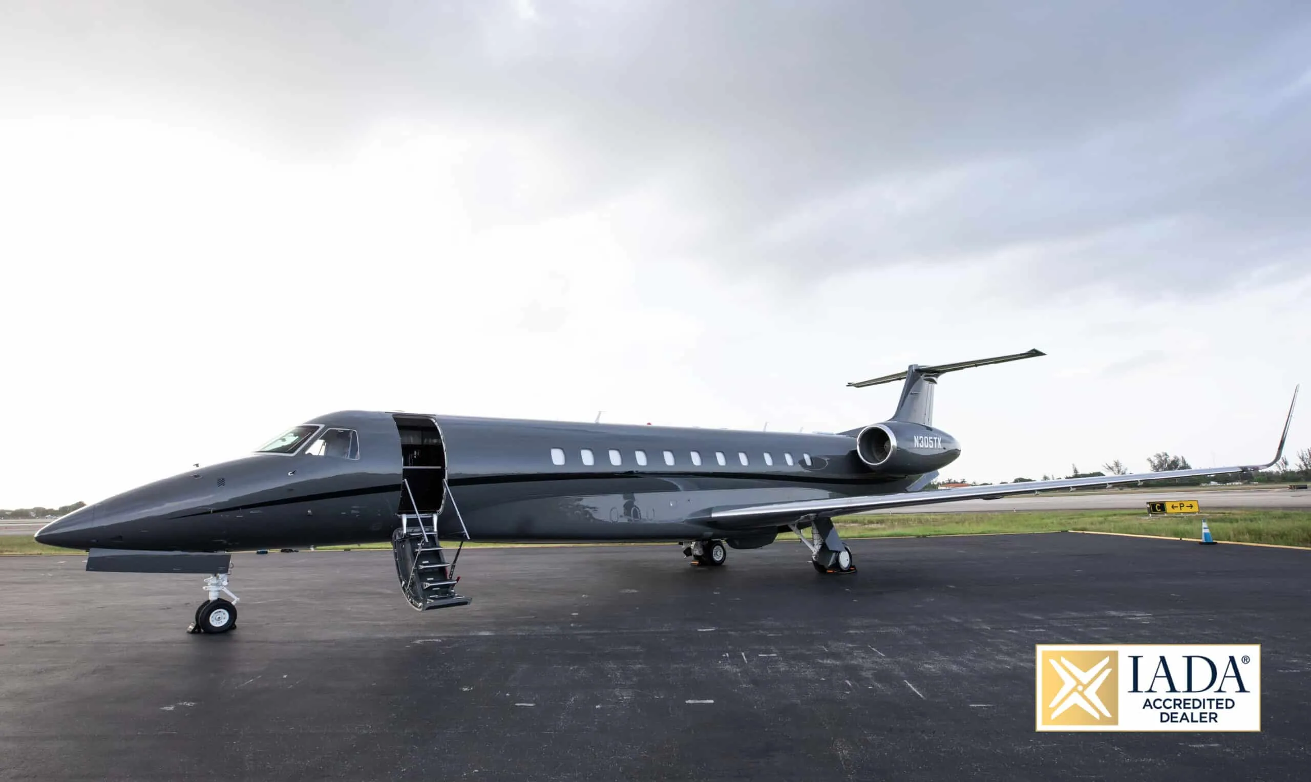A dark grey Embraer Legacy 500 business jet, registration N305TK, parked on a tarmac with its main door open.