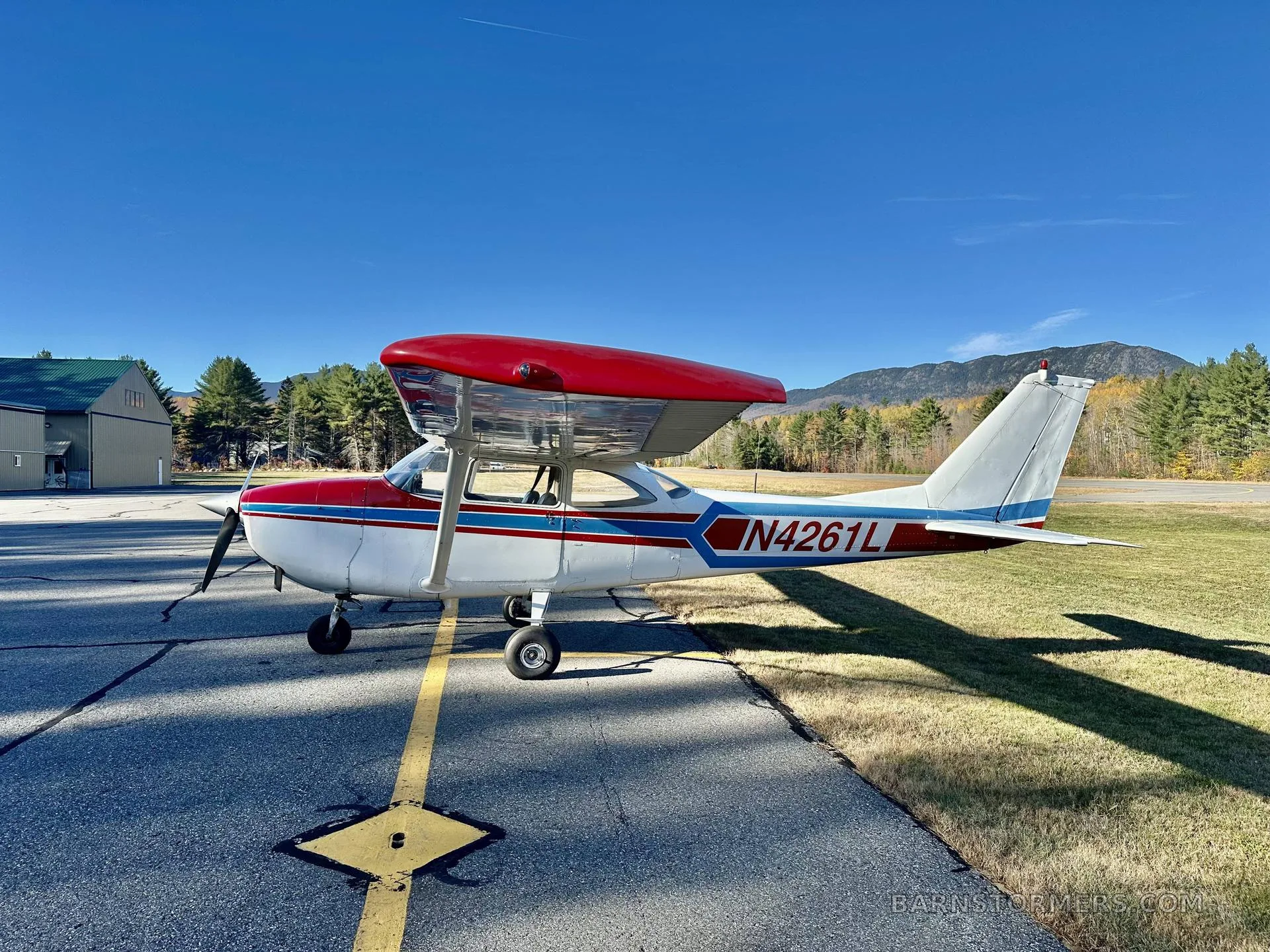 A white single-engine propeller aircraft with red and blue stripes, viewed from the side, parked on an asphalt ramp with a hangar and autumn foliage in the background.