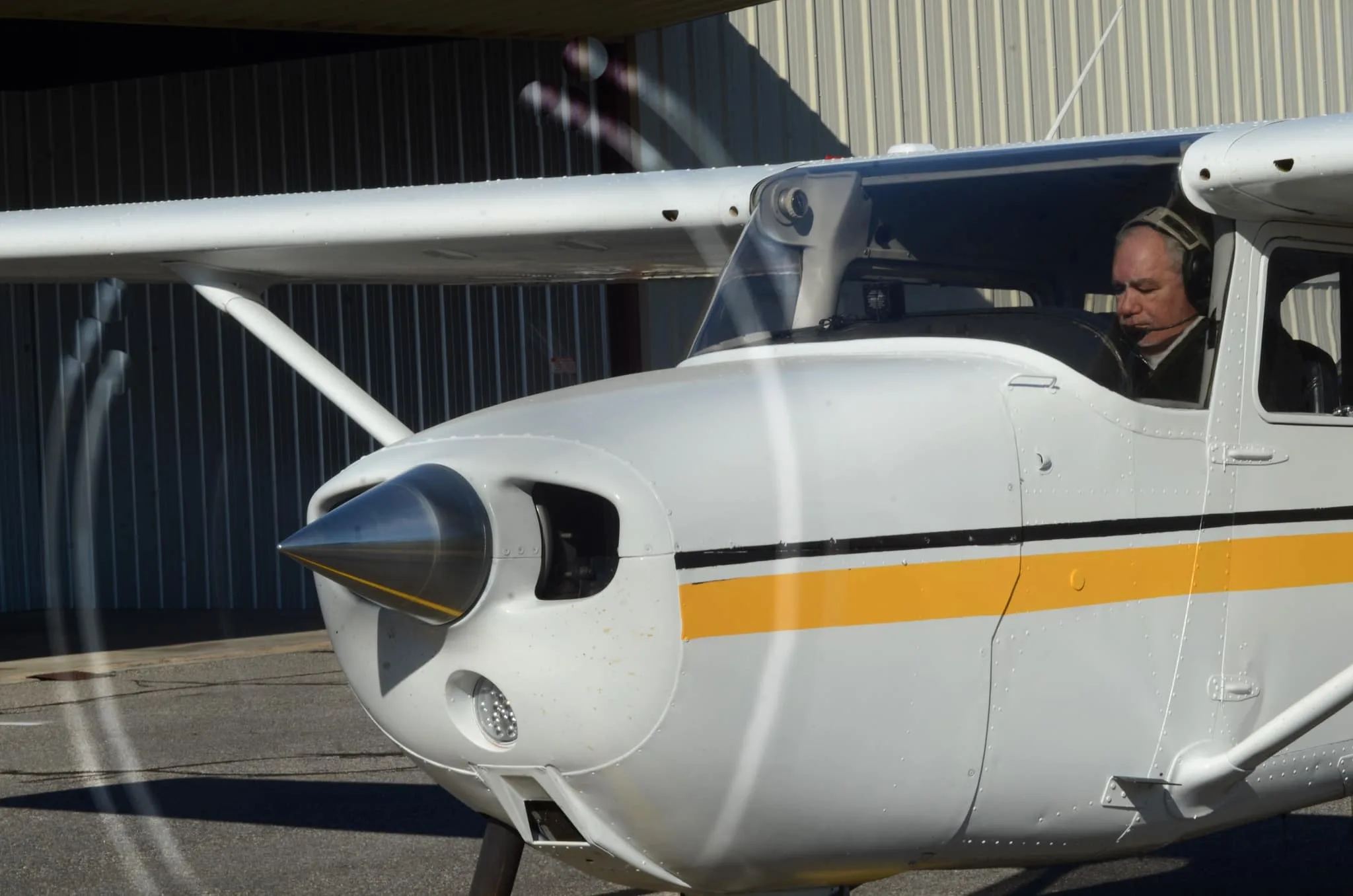 A white high-wing single-engine aircraft with black and yellow stripes, seen from the front-left quarter view on a tarmac with a pilot in the cockpit and a hangar in the background.