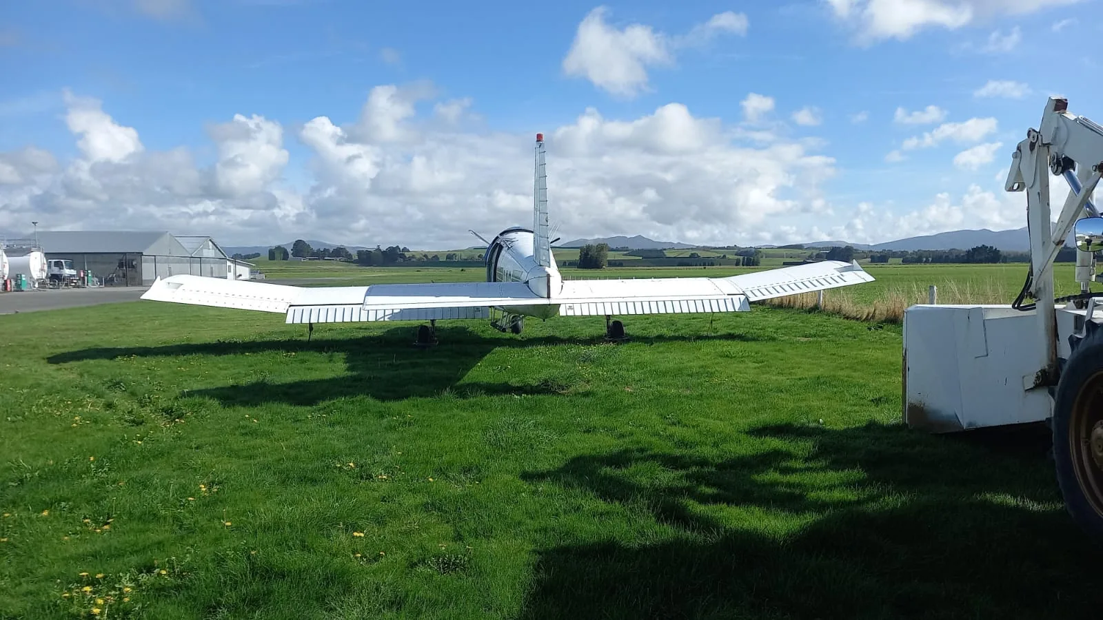 A rear view of a white turboprop aircraft with wide wings, parked on a grassy field with hangars in the distance.