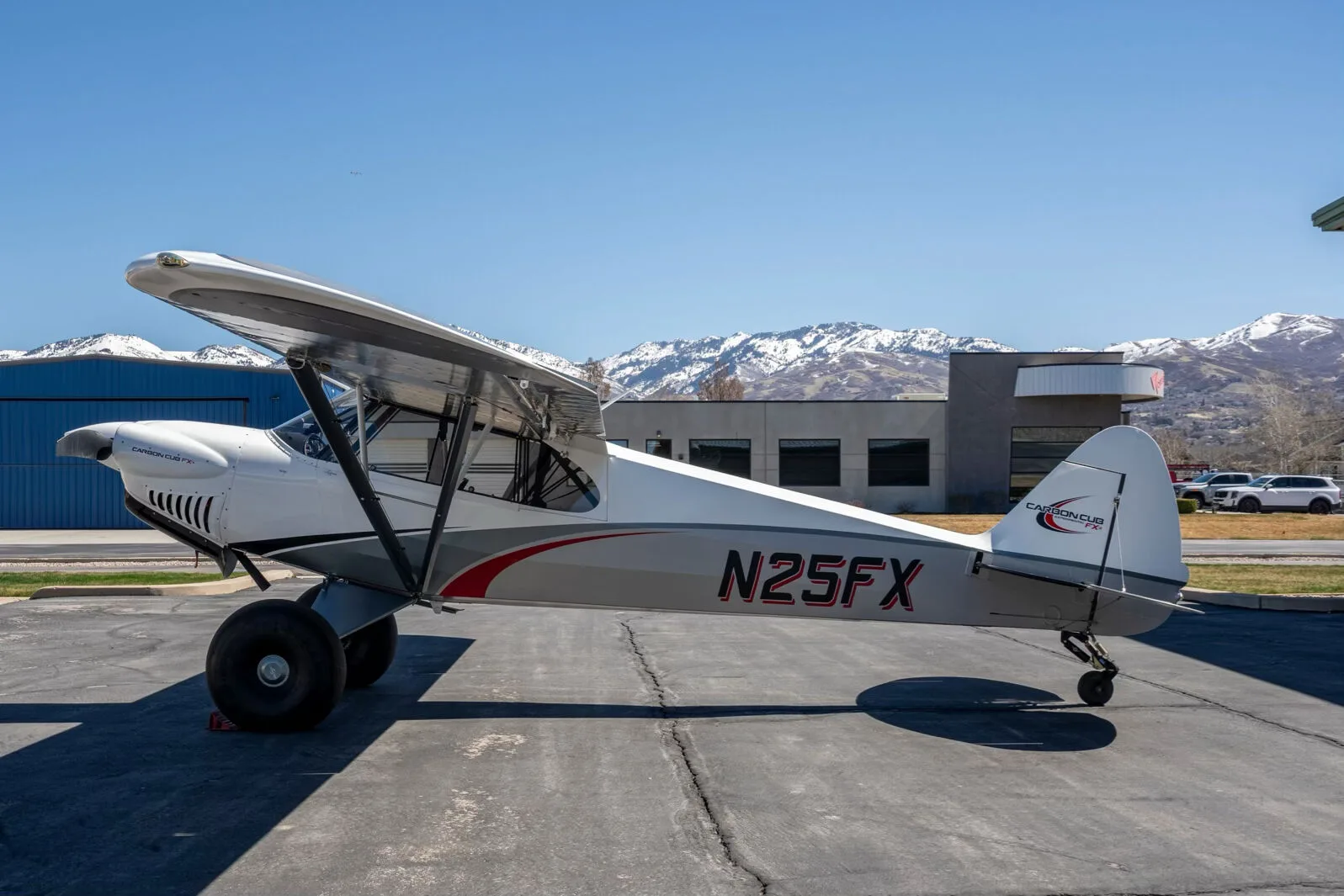 A white and grey Carbon Cub FX-3 aircraft with red accents and registration N25FX parked on an asphalt ramp with snowy mountains in the background.