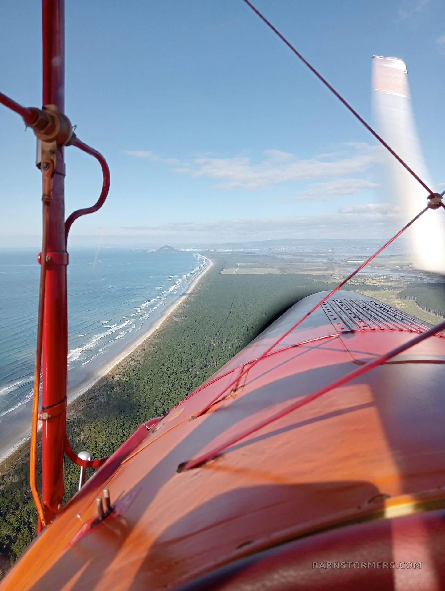 An aerial view from a biplane wing shows a long coastline with a beach, forest, and ocean under a blue sky.