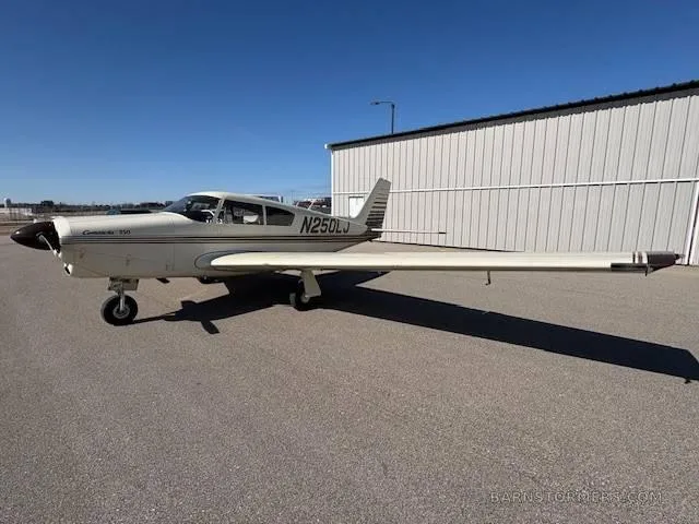 A cream-colored Piper Comanche 250 aircraft with brown stripes, seen from the front-left, parked on an asphalt apron next to a hangar.