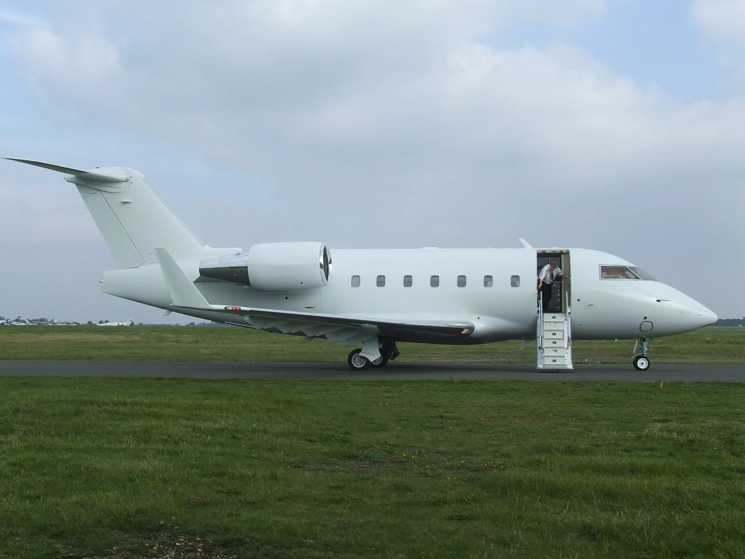 A white Bombardier Challenger business jet with its main door open and a person standing inside, parked on a grassy airfield.