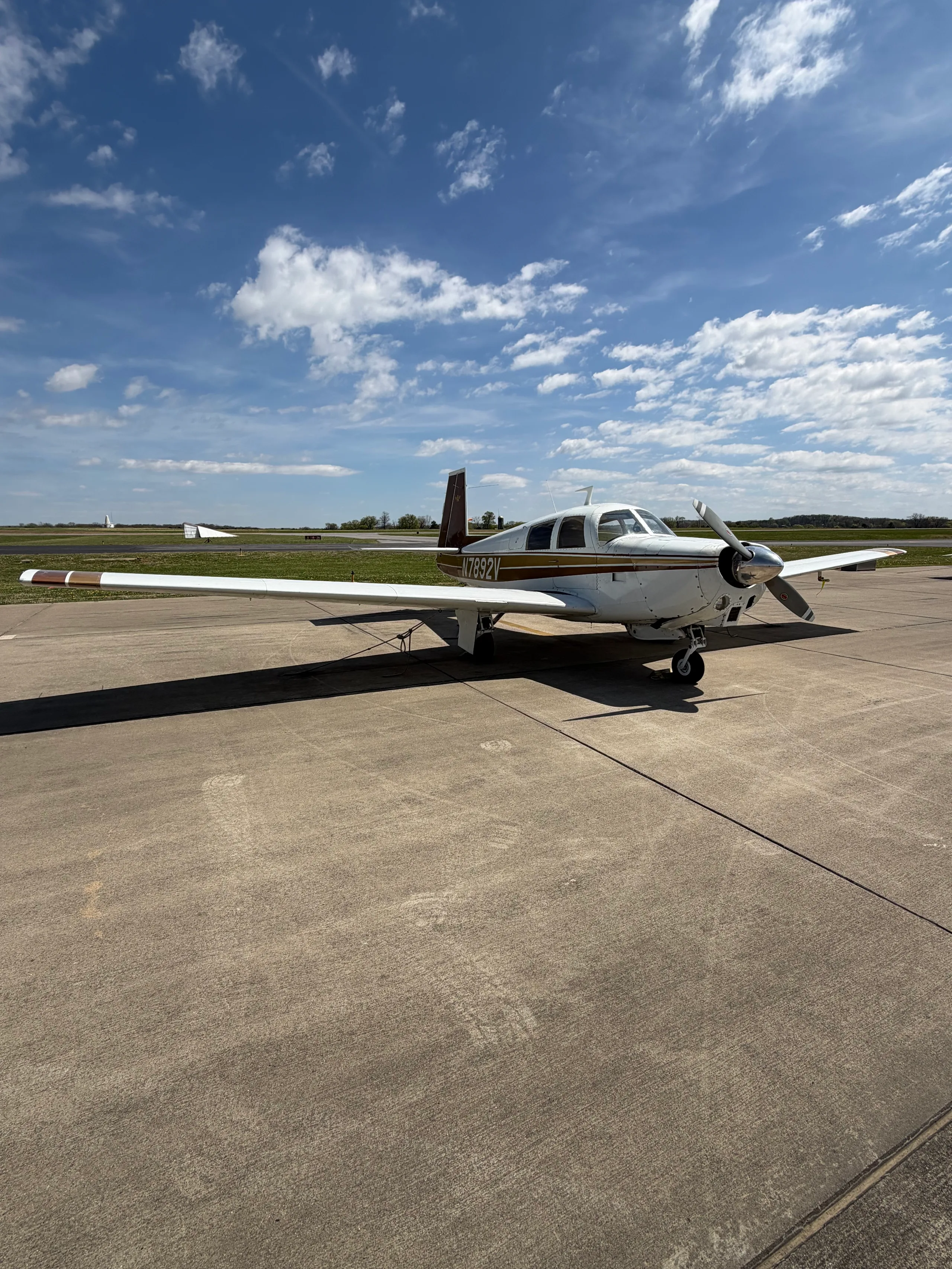 A white and brown single-engine aircraft (N7892V) with its propeller visible, parked on an airport tarmac.
