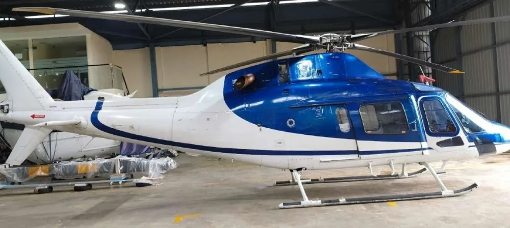 A three-quarter view of a blue and white helicopter with a white tail, parked in a hangar with other equipment.