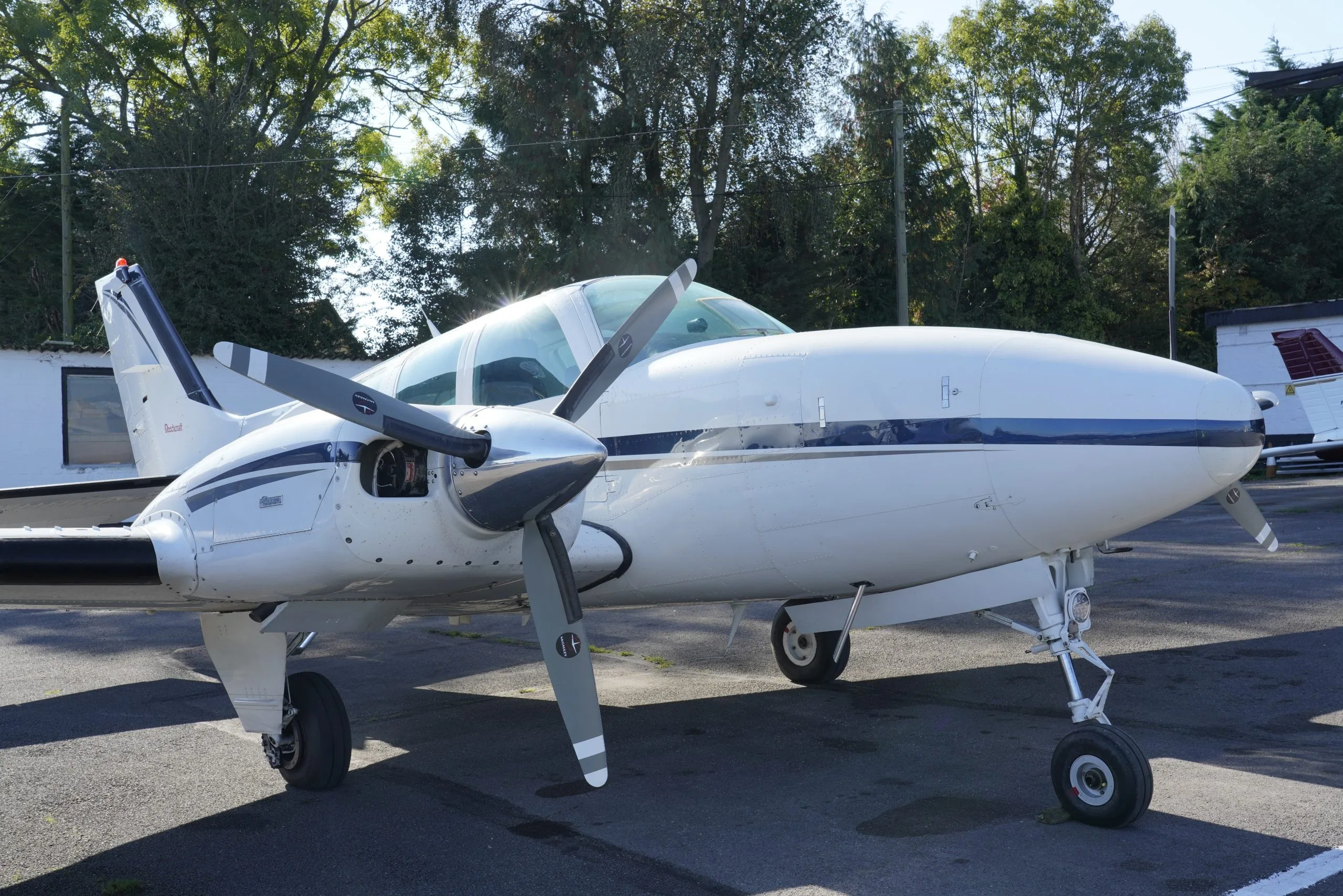 A front-side view of a white twin-engine propeller aircraft with blue stripes, parked on an asphalt apron.