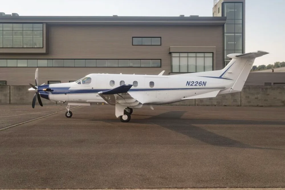 A white and blue Pilatus PC-12 aircraft with registration N226N parked on an tarmac next to a modern building.