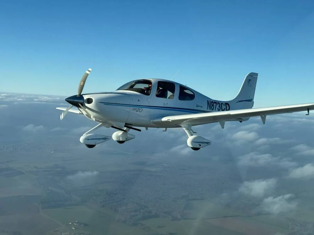 A white Cirrus SR20 aircraft (N873CD) with blue stripes flying above clouds and green landscape.