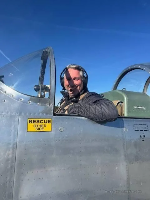 A smiling pilot in a headset looks out from the cockpit of a silver experimental aircraft under a clear blue sky.