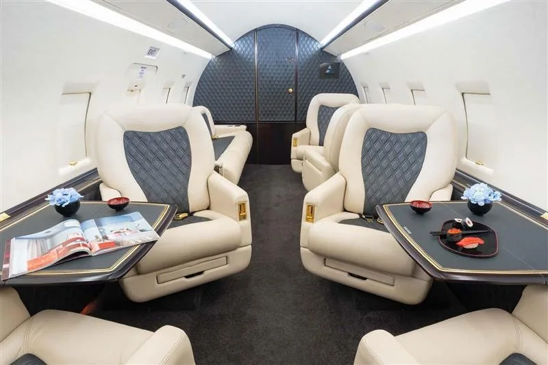 A luxurious private jet cabin with cream and dark grey quilted seats and tables set with sushi and a magazine.