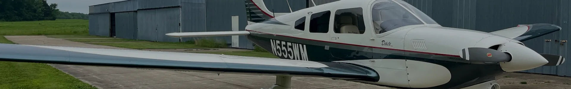 A white and dark green single-engine propeller aircraft, possibly a Piper Dakota, with registration N55WM, parked on an apron in front of a hangar.