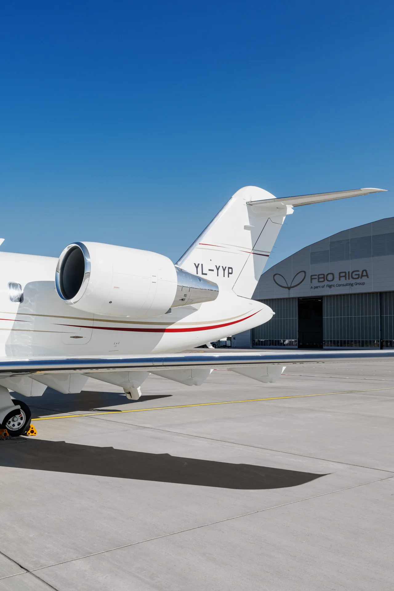 The tail section and engine of a white Bombardier Challenger 605 business jet, registration YL-YYP, parked on a tarmac with an FBO RIGA hangar in the background.