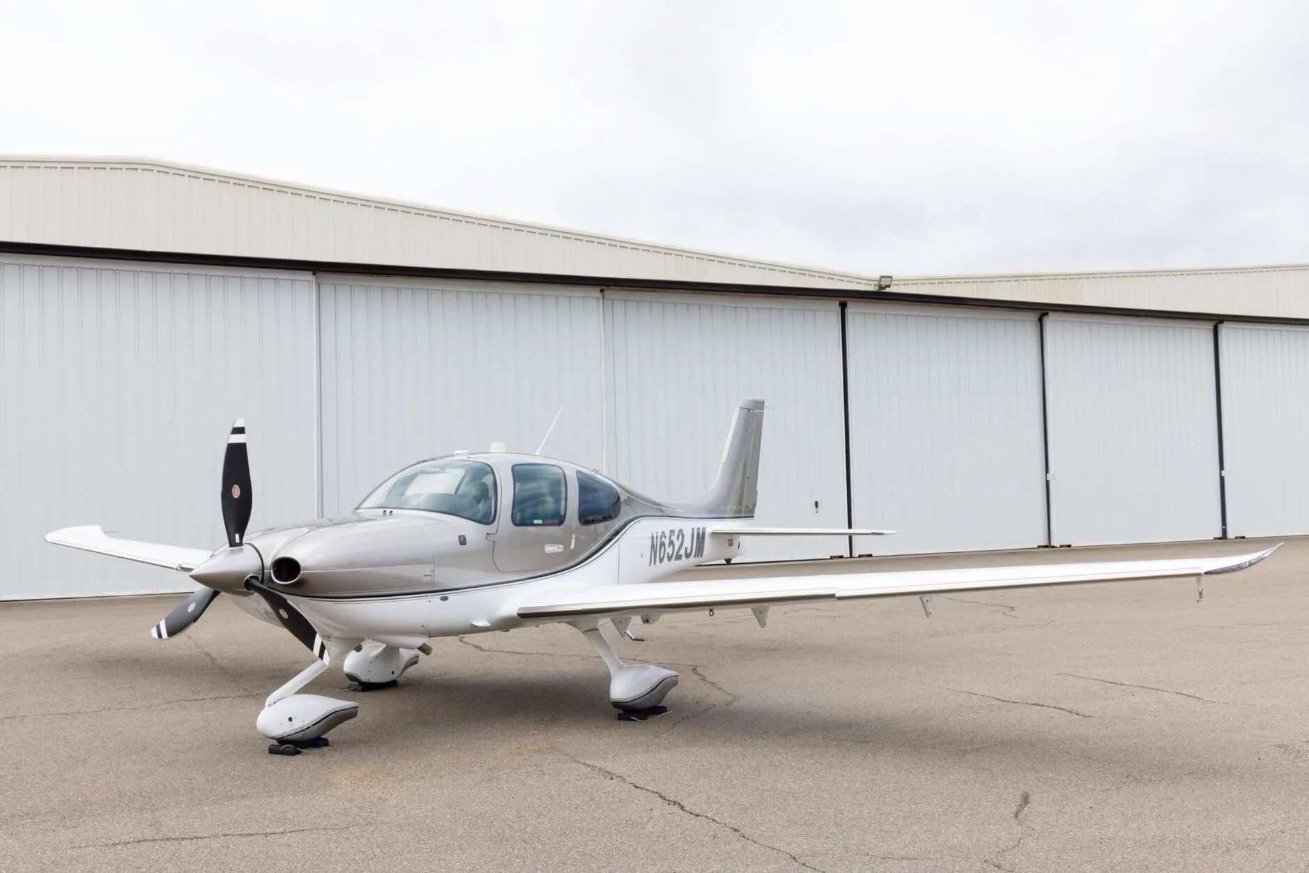 A Cirrus SR22 aircraft with N652JM registration, painted in silver and white, parked on an tarmac in front of hangars.