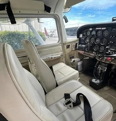 The light-colored interior cabin seats and partial cockpit of a small aircraft, with another aircraft visible outside.