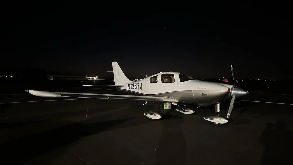 A white and silver Columbia 350 aircraft, N1267J, parked on an airport tarmac at night under artificial lighting.