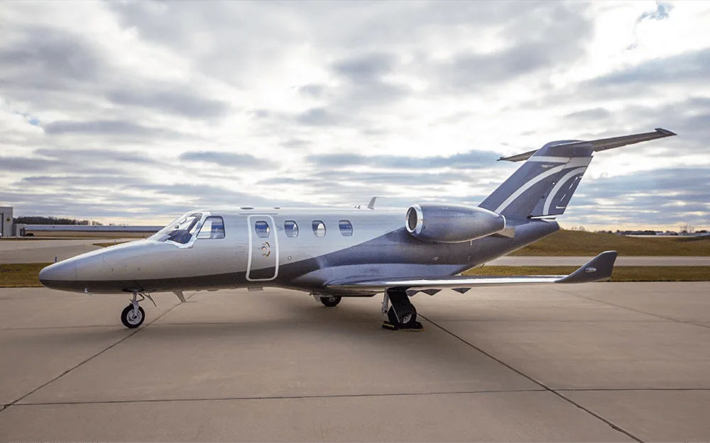 A silver and dark grey business jet is parked on an asphalt tarmac under a cloudy sky.