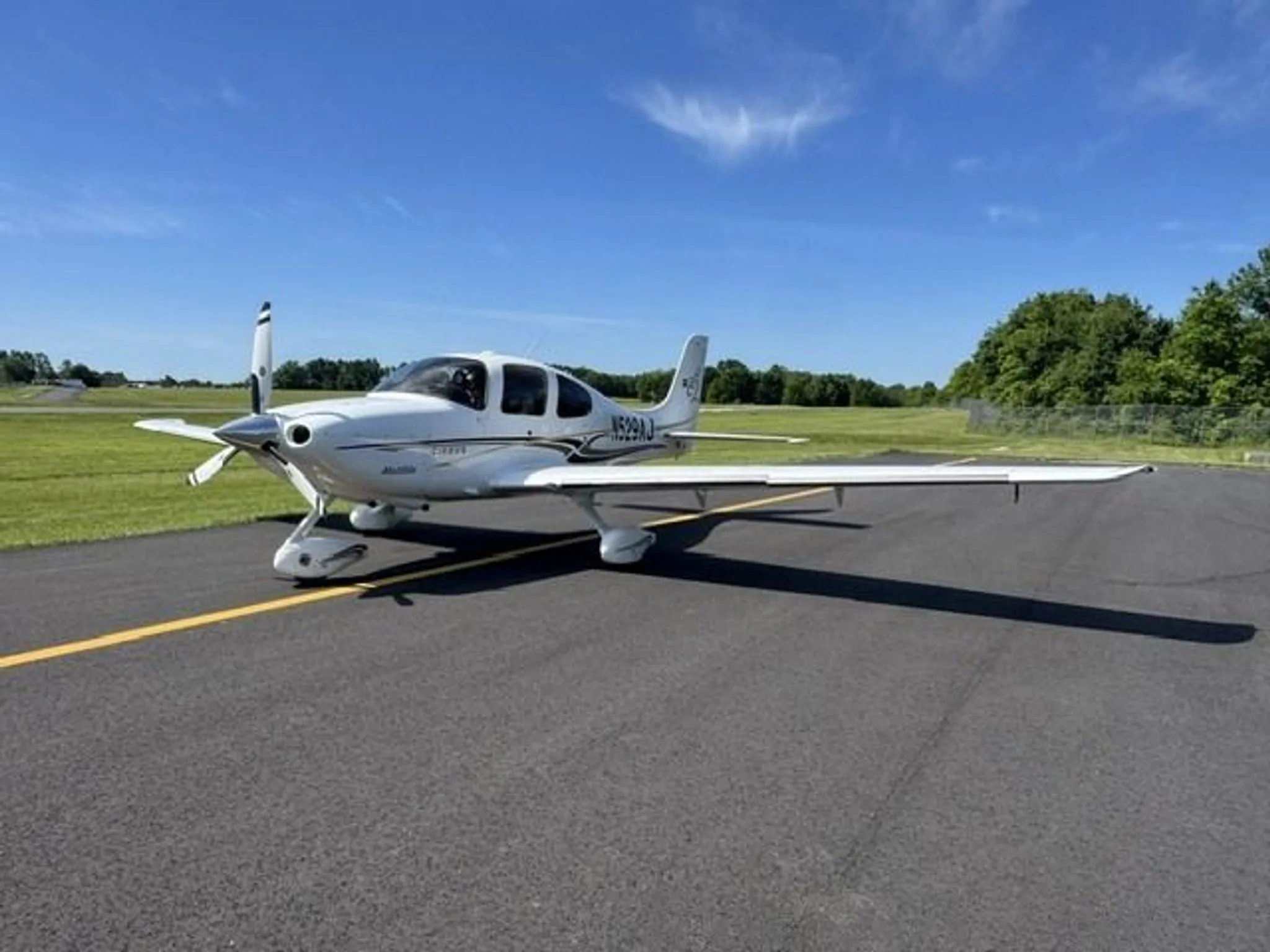 A white Cirrus SR22 aircraft with registration N529AJ parked on an asphalt surface under a clear blue sky.