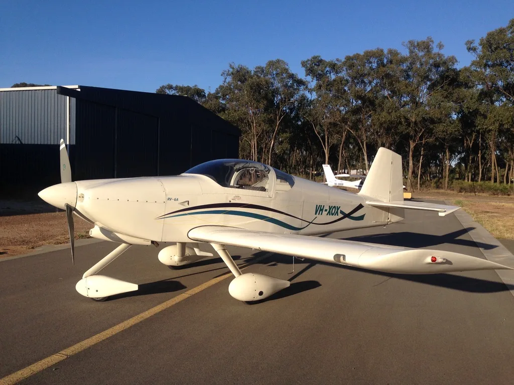 A white RV-6A aircraft with blue and black stripes is parked on an asphalt runway next to a hangar, with trees in the background.