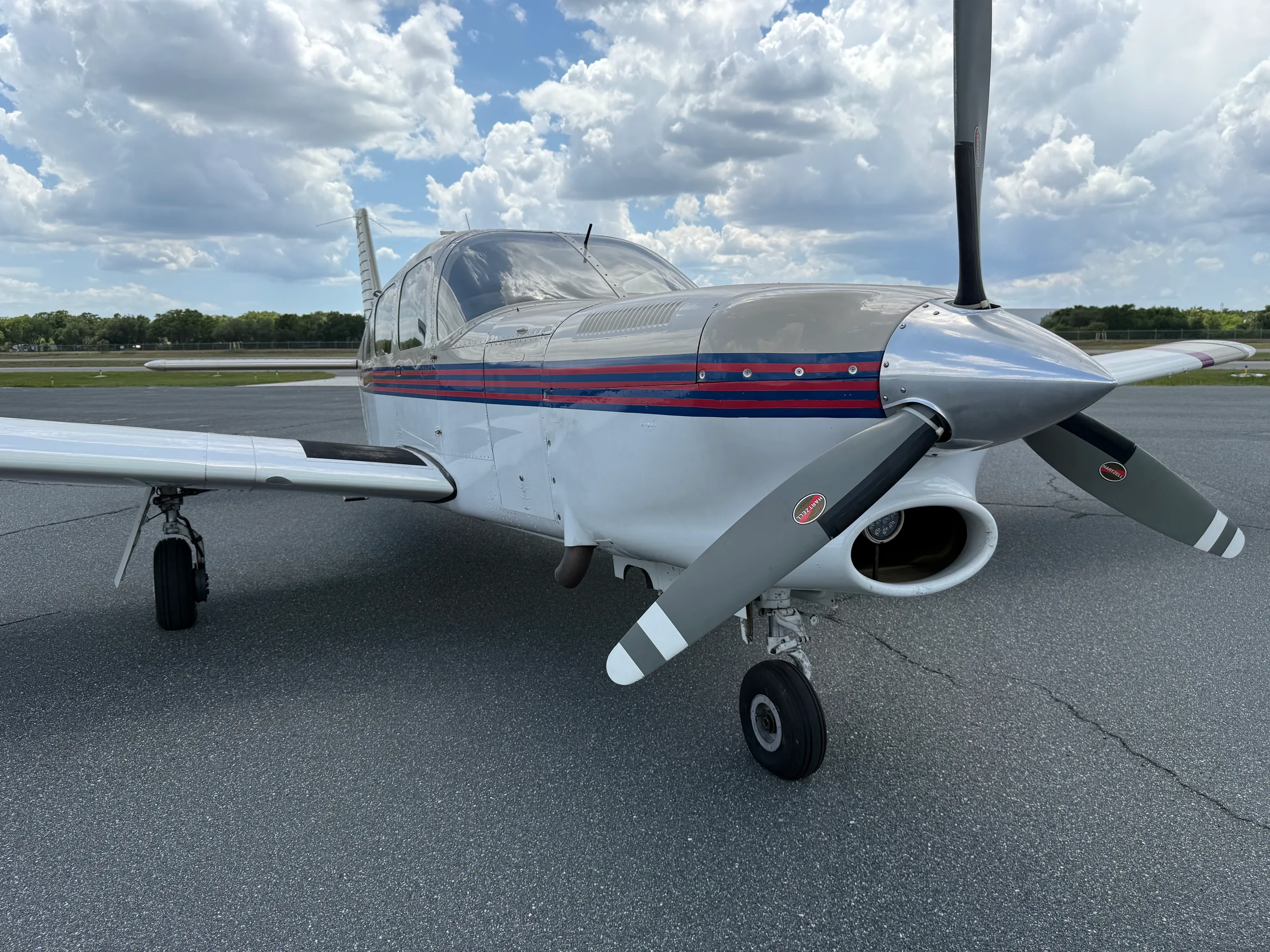 A white and grey single-engine propeller aircraft with red and blue stripes is parked on an asphalt tarmac under a cloudy sky.